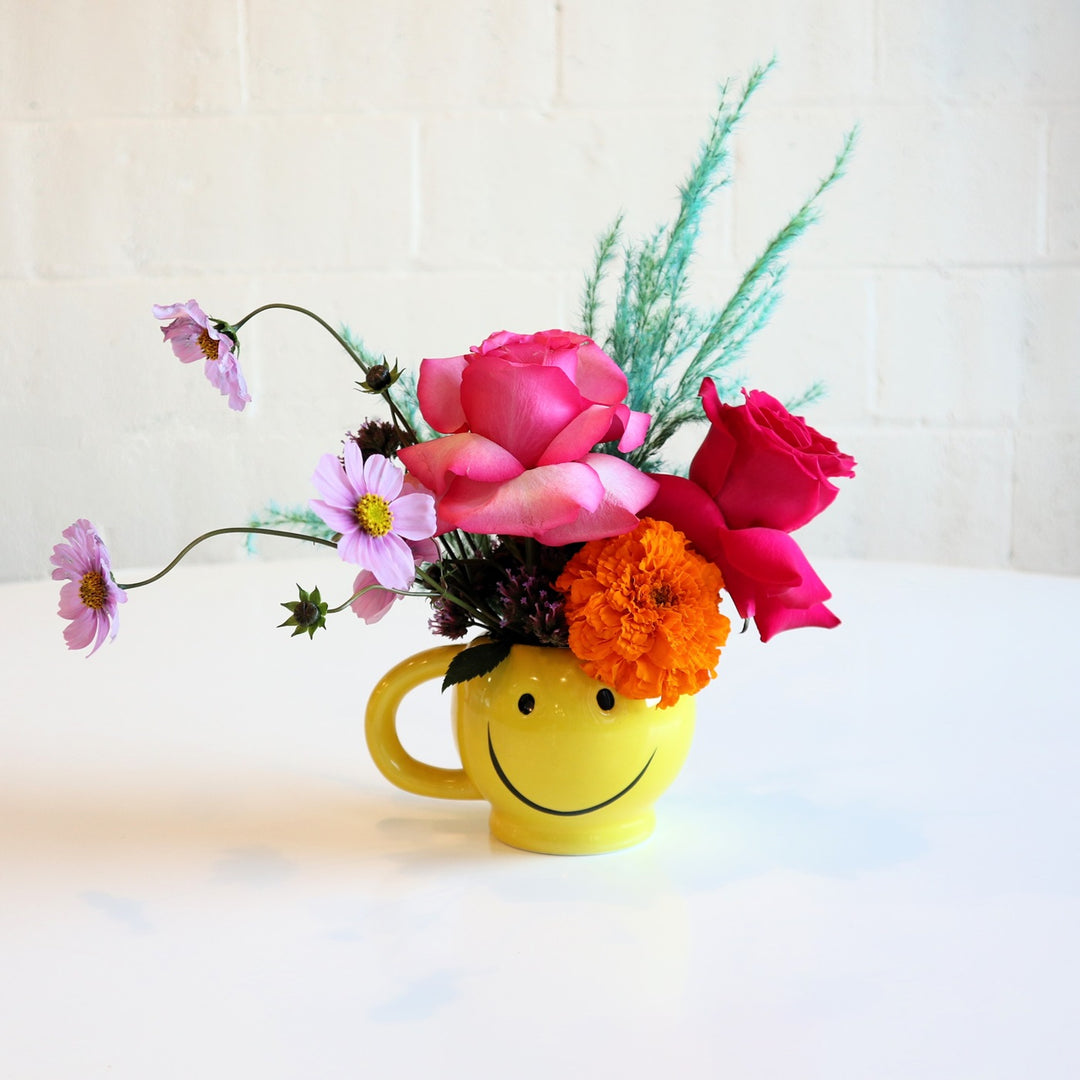 Smiley Mug | A bold yellow smiley mug with an arrangement of purple, pink, orange, and blue florals. Photo taken against a white background.