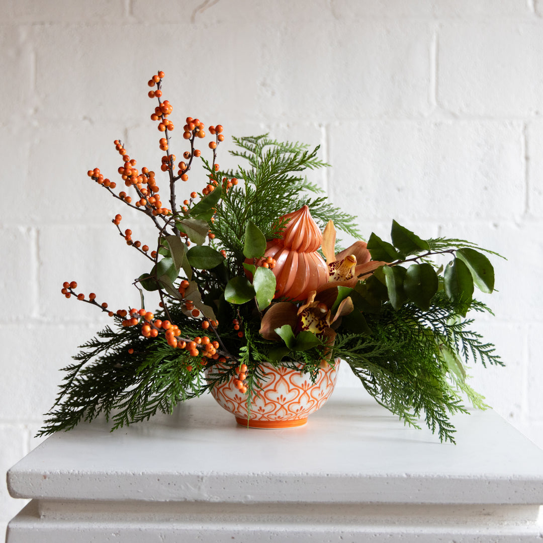 Decorative arrangement with greenery, berries, and a candle in an orange holder on a white surface.