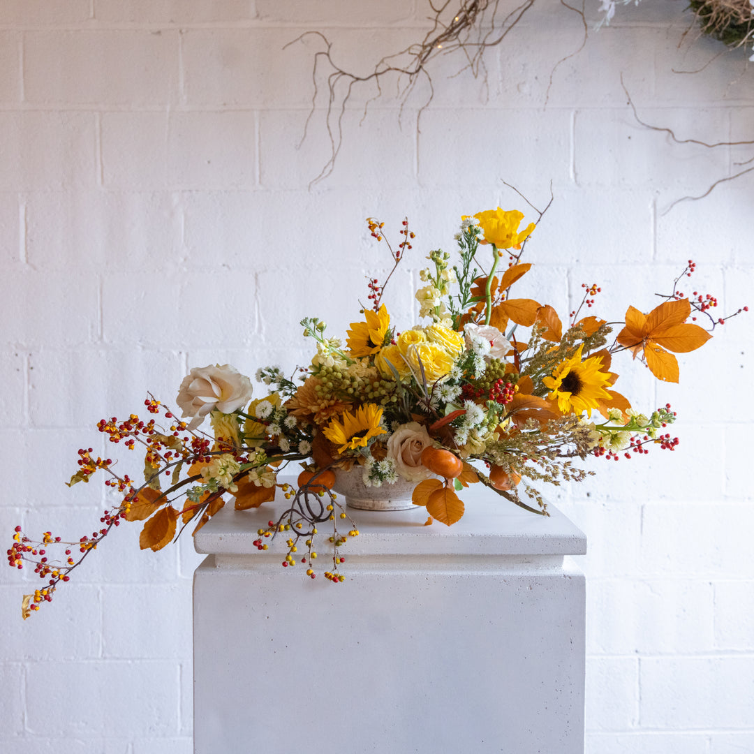 Autumnal floral arrangement with yellow and orange flowers on a white pedestal against a white brick wall.