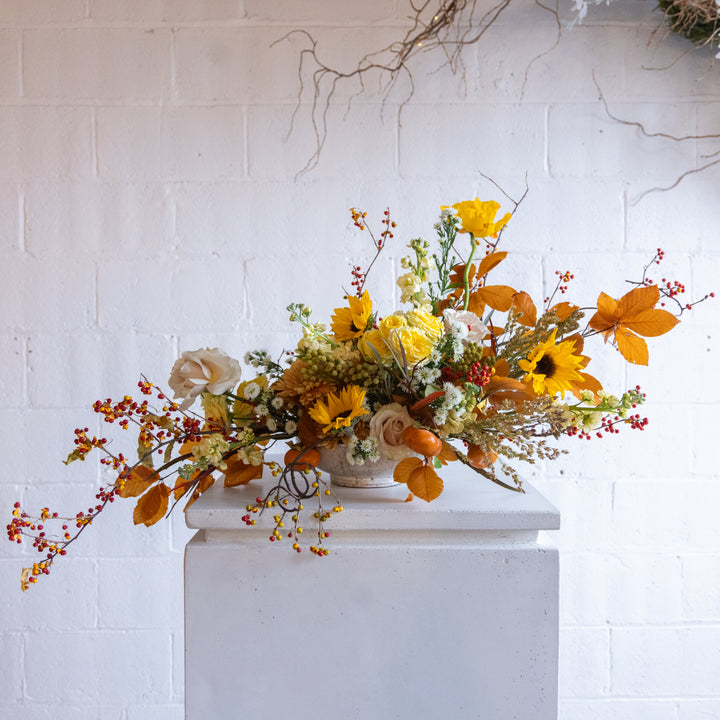 Autumnal floral arrangement with yellow and orange flowers on a white pedestal against a white brick wall.