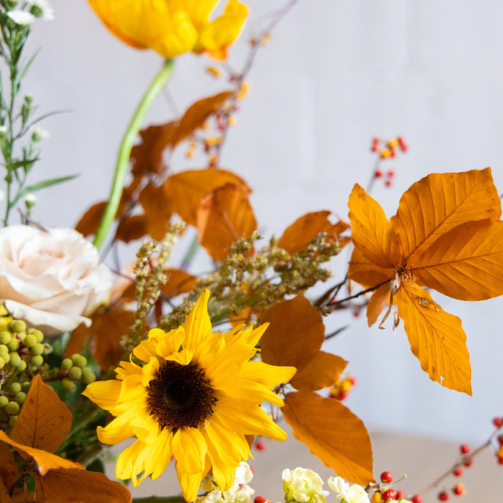 Autumn-themed floral arrangement with sunflowers, roses, and leaves on a light background