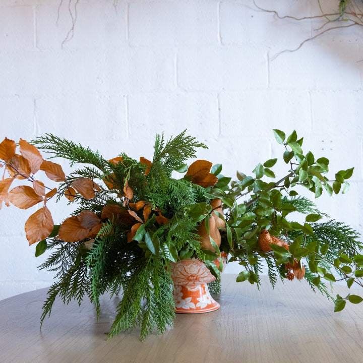 Decorative arrangement of green and brown leaves in a vase on a light surface with a white textured background.