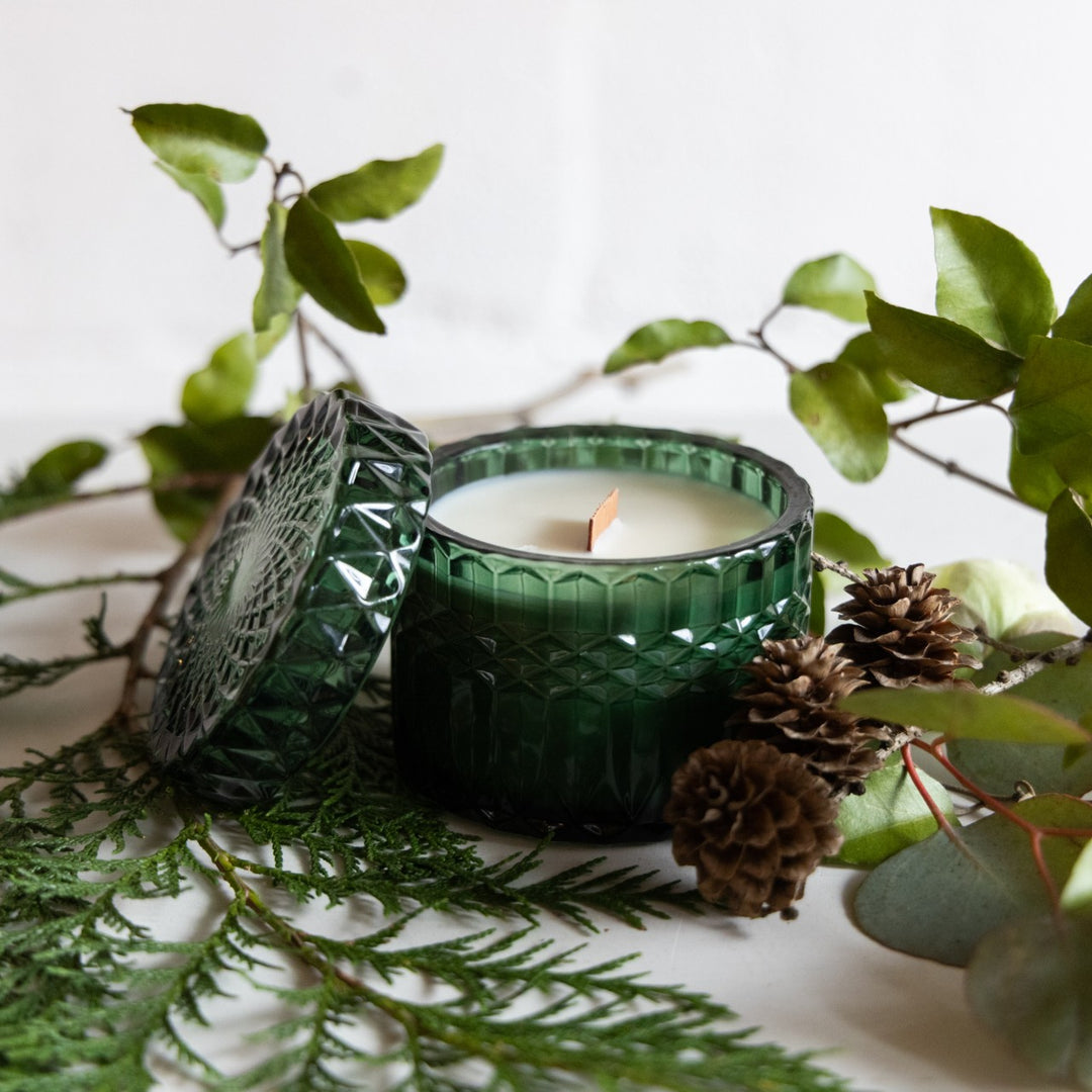 Green textured candle holder with a lit candle surrounded by greenery and pinecones on a light background