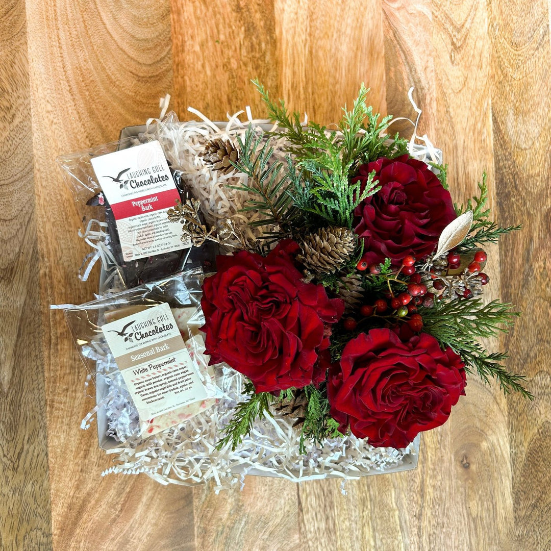 A holiday gift box featuring a vibrant floral arrangement with deep red roses, lush evergreen sprigs, pinecones, and red berries. The box also includes gourmet treats, such as Peppermint Bark and White Peppermint Bark from Laughing Gull Chocolates, nestled in natural shredded filler for a festive presentation. Perfect for gifting or holiday decor.