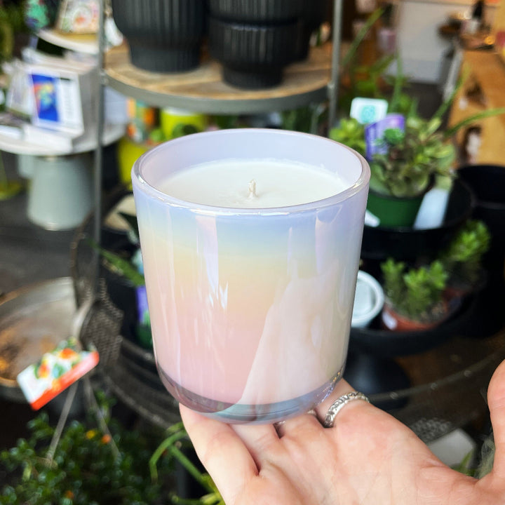 A hand holding a glass jar candle with a pastel iridescent gradient from lavender to pink and cream, set against a blurred shop background with plants and pots.