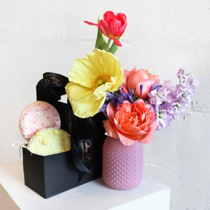 A black carrier filled with an assortment of gourmet goodies baked cookies and vibrant, fragrant flowers, offering a delightful combination of sweet treats and natural beauty.