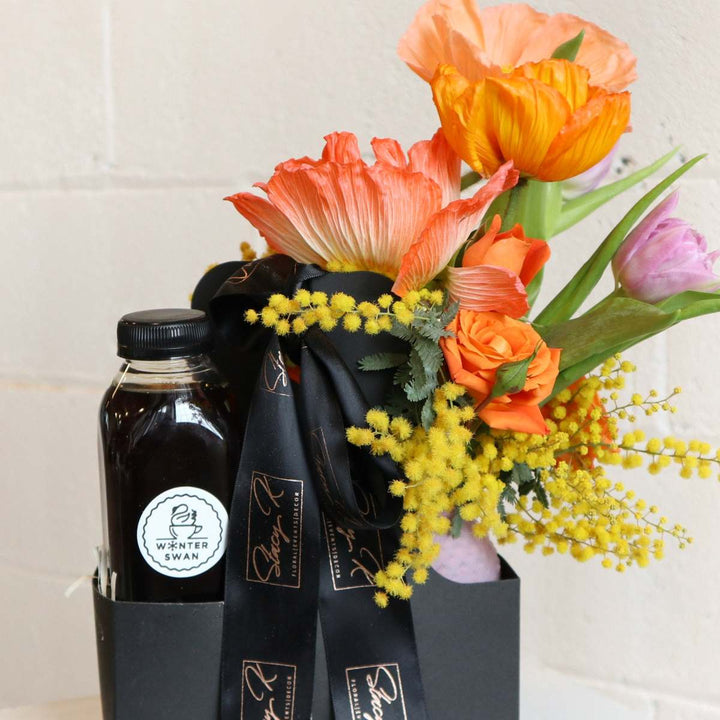 Gift box featuring vibrant orange poppies, tulips, mimosa, and roses arranged alongside a bottle of premium cold brew coffee, wrapped with a black ribbon.