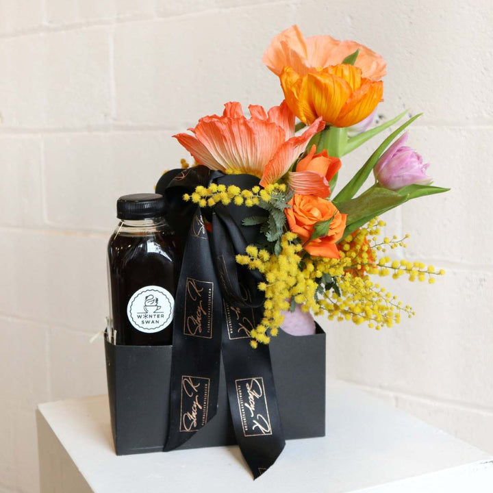 Gift box featuring vibrant orange poppies, tulips, mimosa, and roses arranged alongside a bottle of premium winter swan cold brew coffee, wrapped with a black ribbon.