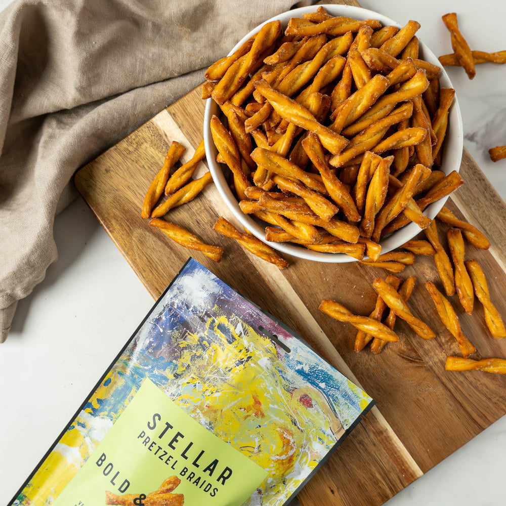 Bowl of pretzel sticks on a wooden board with a package labeled 'Stellar Pretzel Breads' on a light background.
