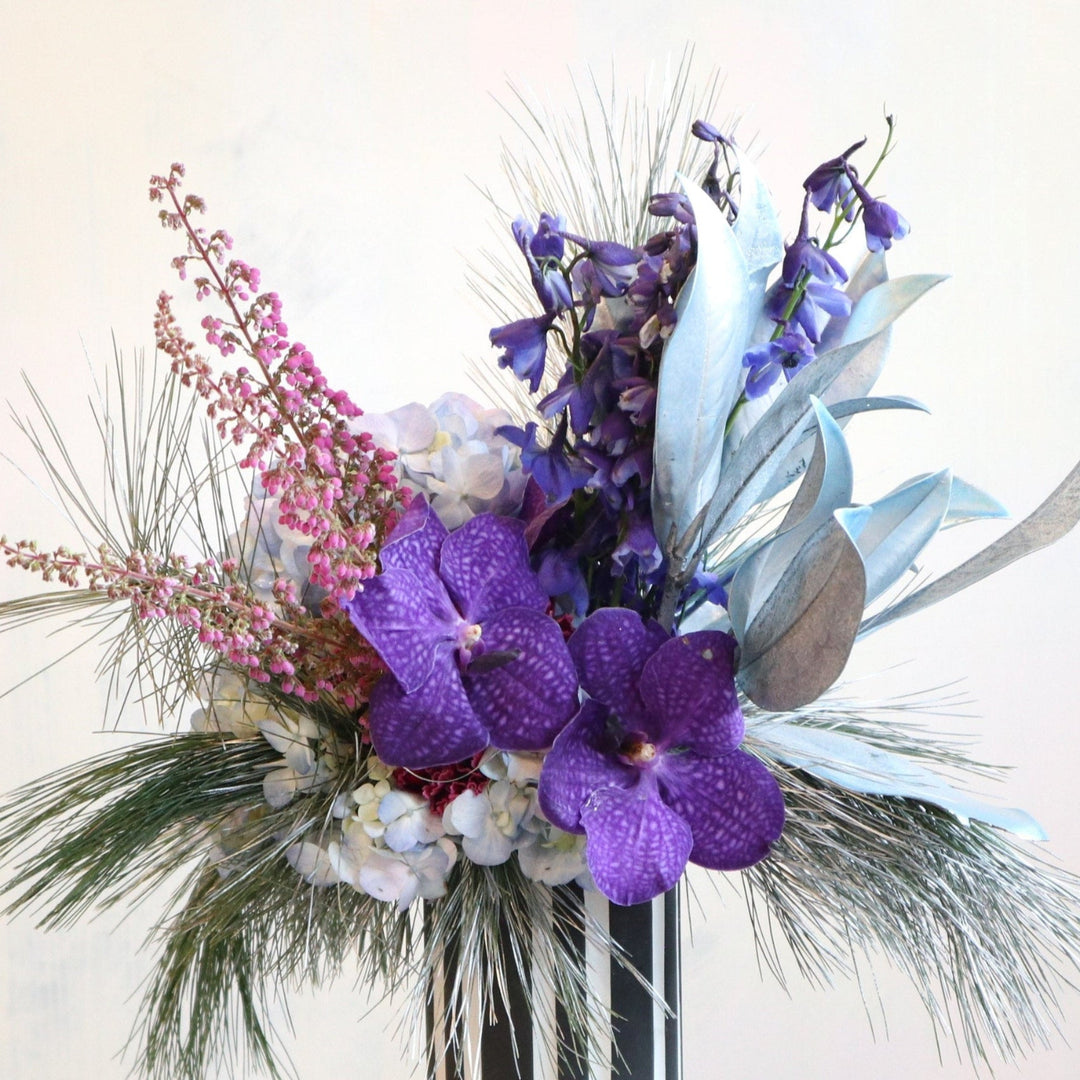 Vased flower arrangement, purple vanda orchids, painted magnolia, blue delphinium, hydrangea, silver white pine. 