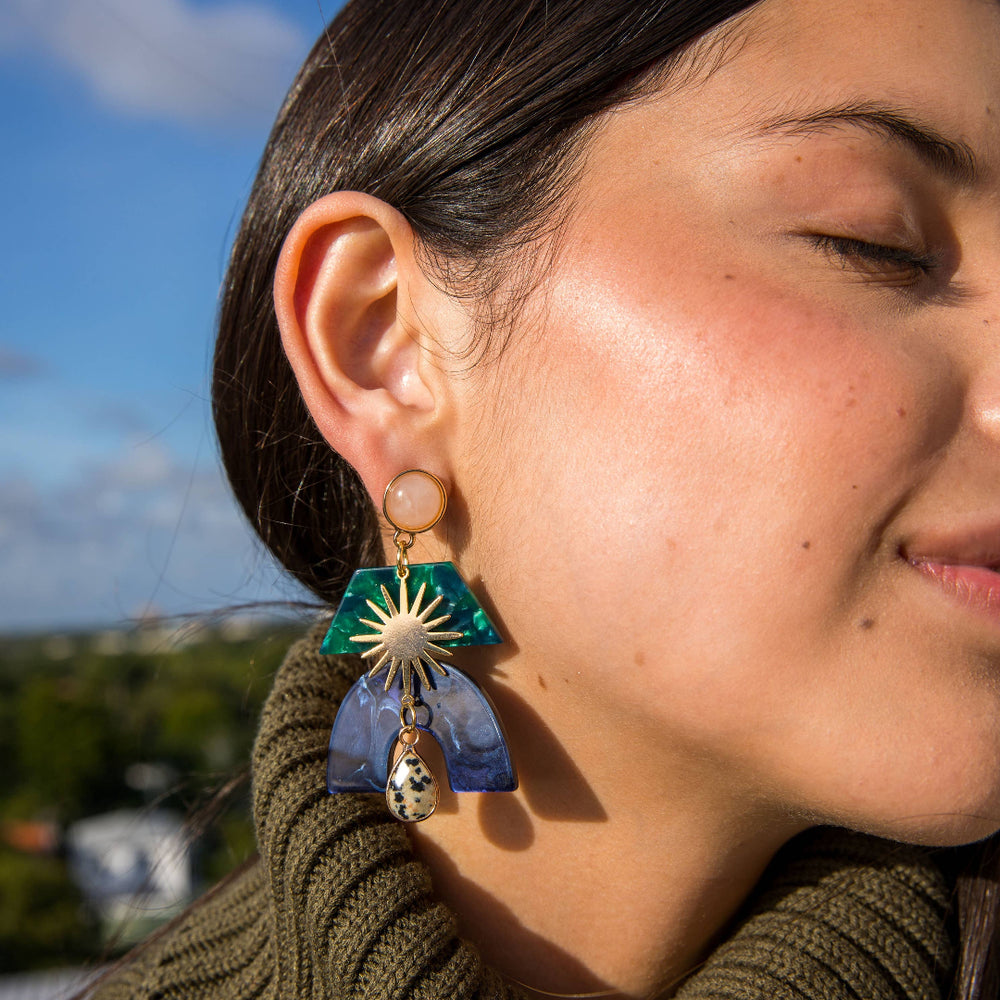 Close-up of a person wearing a unique earring with a blue and green design, set against a blurred outdoor background.