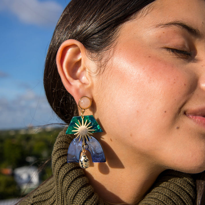 Close-up of a person wearing a unique earring with a blue and green design, set against a blurred outdoor background.