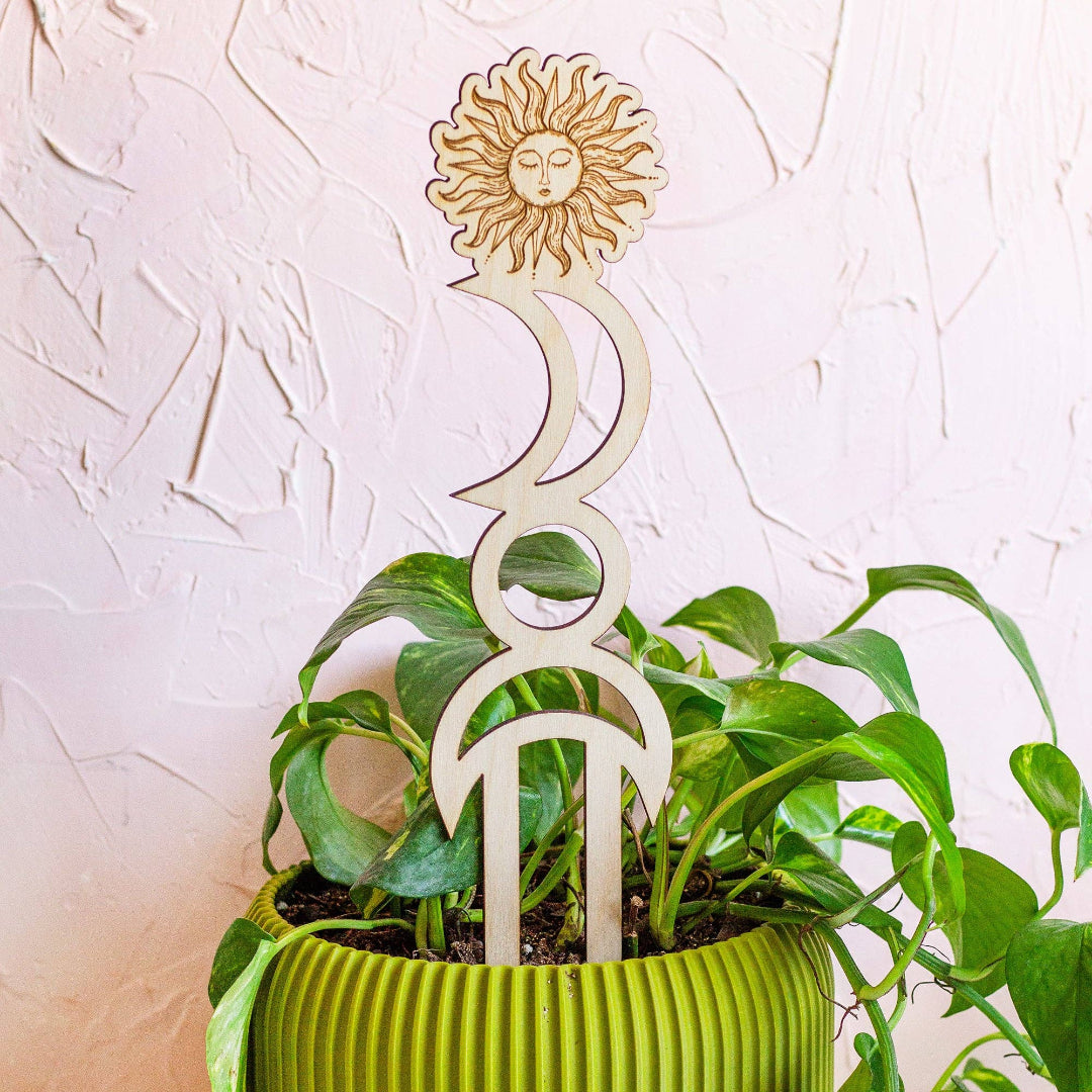 Decorative plant stake with sun and moon design in a pot against a textured white wall.