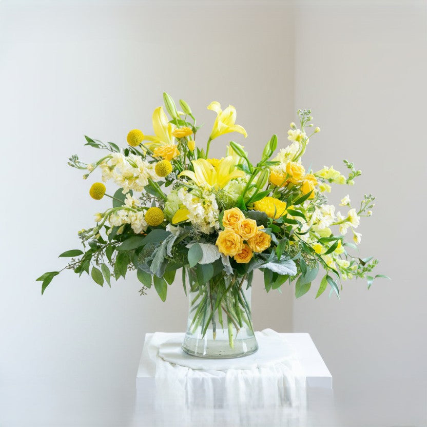 Bouquet of yellow and white flowers in a clear vase on a white surface with a light gray background