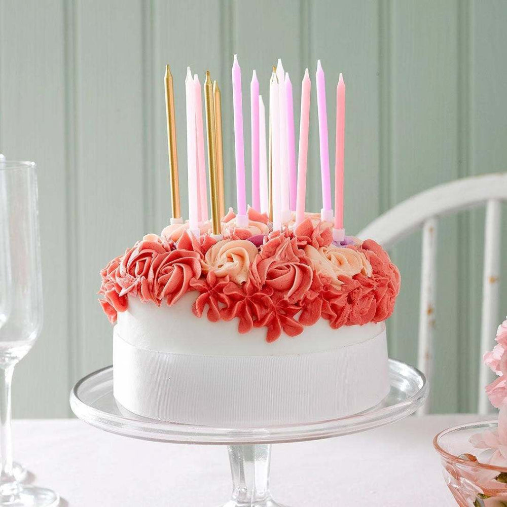 Talking Tables | Pink and gold cake candles | White cake with pink and gold candles on a clear cake stand against a light green wall.