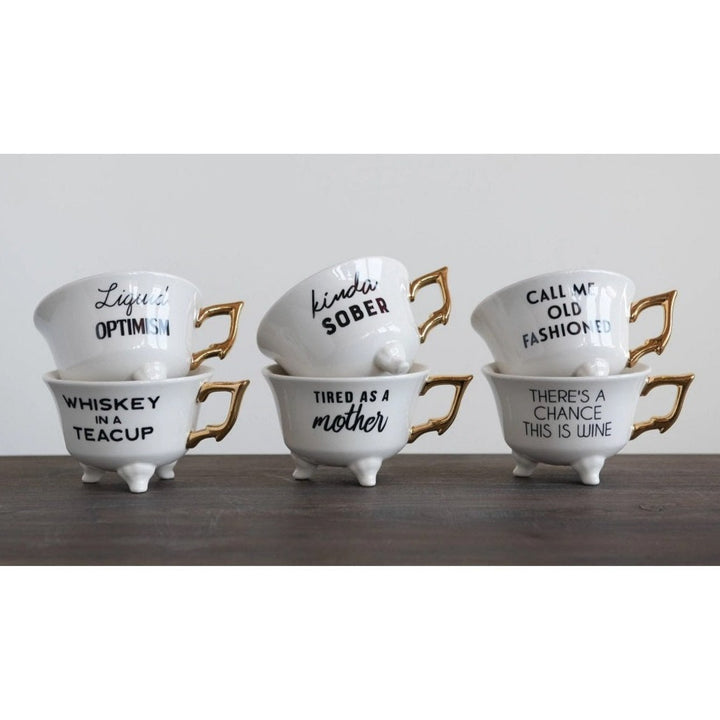 Saying Teacups | White teacups black text little feet and gold handles. Each cup has a different saying: "Liquid Optimism" "Whiskey In A Teacup" "Kinda Sober" "Tired As A Mother" "Call Me Old Fashioned" "There's A Chance This is Wine".