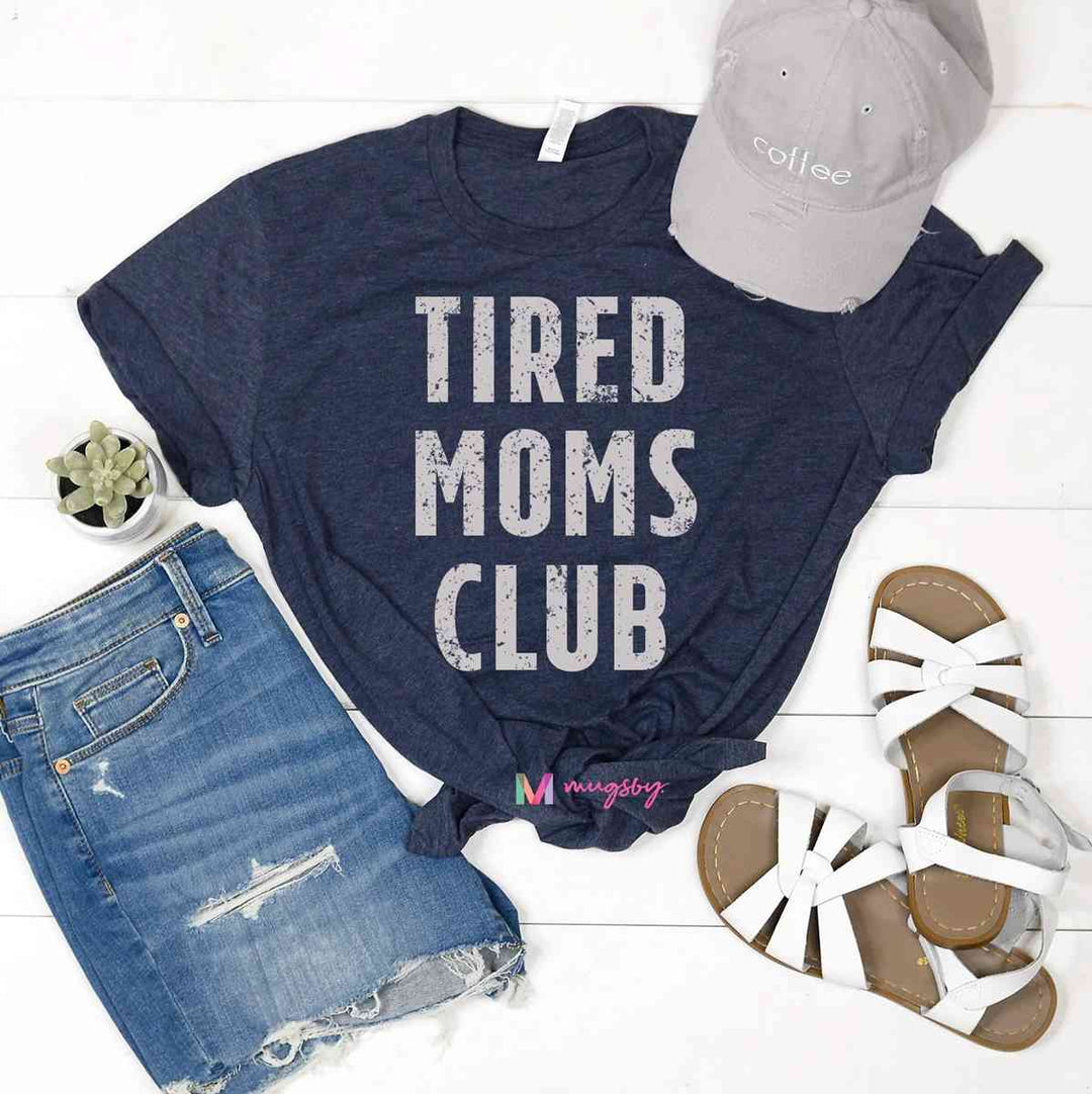 Mugsby | Tired Moms Club | A gray/blue tshirt with bold text reading tired moms club. A succulent, jean shorts, baseball cap, and shoes are used to stage the photo.