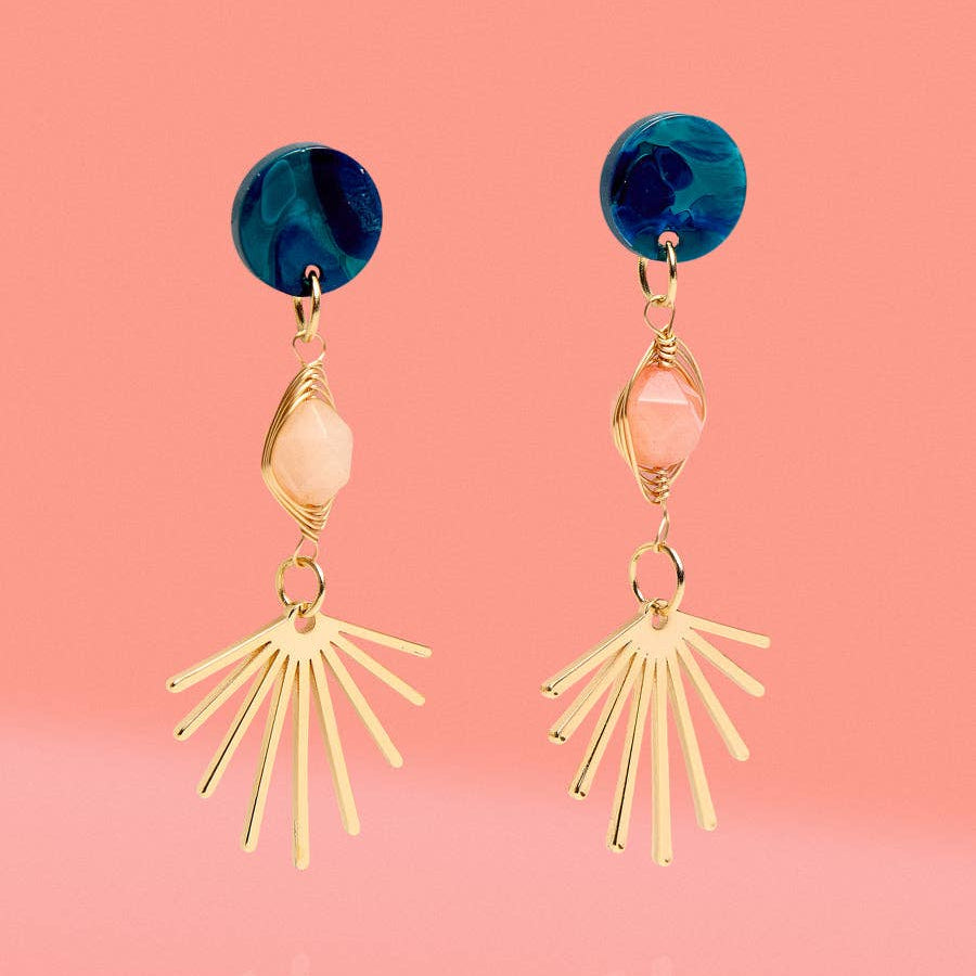 A pair of gold-toned dangle earrings with a deep blue round stud, a pale pink faceted bead, and a fan-like burst of thin gold bars, shown on a pink gradient background.