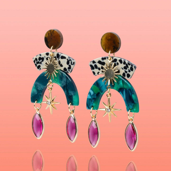 A pair of bohemian-style drop earrings featuring a brown jasper stone, a black-and-white leopard print band, a teal and green marbled arch, gold starburst charms, and dangling magenta teardrop gemstones, set against a pink gradient background.