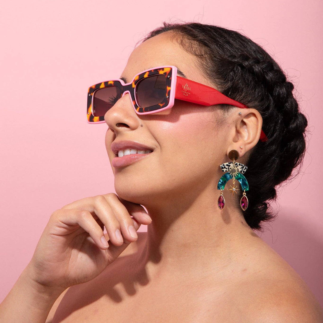 A woman with dark braided hair wearing square sunglasses with a pink and tortoiseshell frame, posed against a solid pink background.