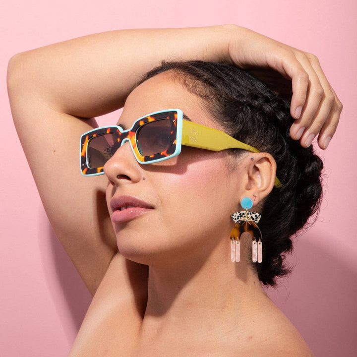A woman with dark hair in a half-up style wearing colorful square sunglasses with a yellow-green frame and tortoiseshell accents, posed against a solid pink background.