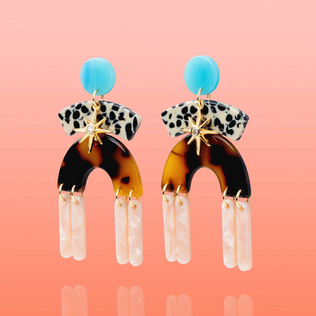 A pair of statement drop earrings on a peach-pink gradient background. The earrings feature a bright blue circular top, a black-and-white leopard print band, a gold starburst connector, a large tortoiseshell arch, and three dangling white textured bar charms.
