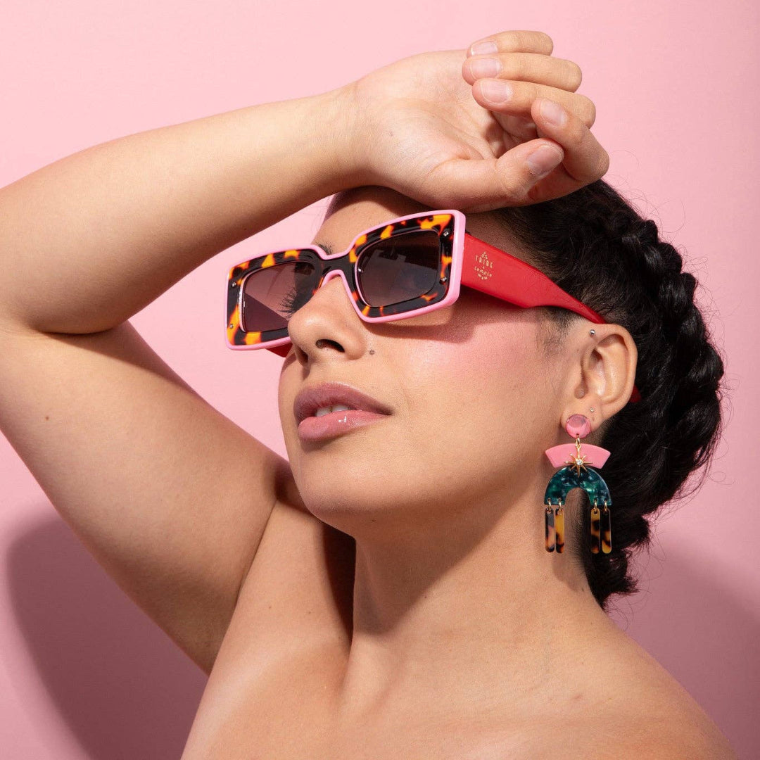 A woman wearing pink and tortoiseshell patterned sunglasses with red temple arms, posing against a solid pink background.