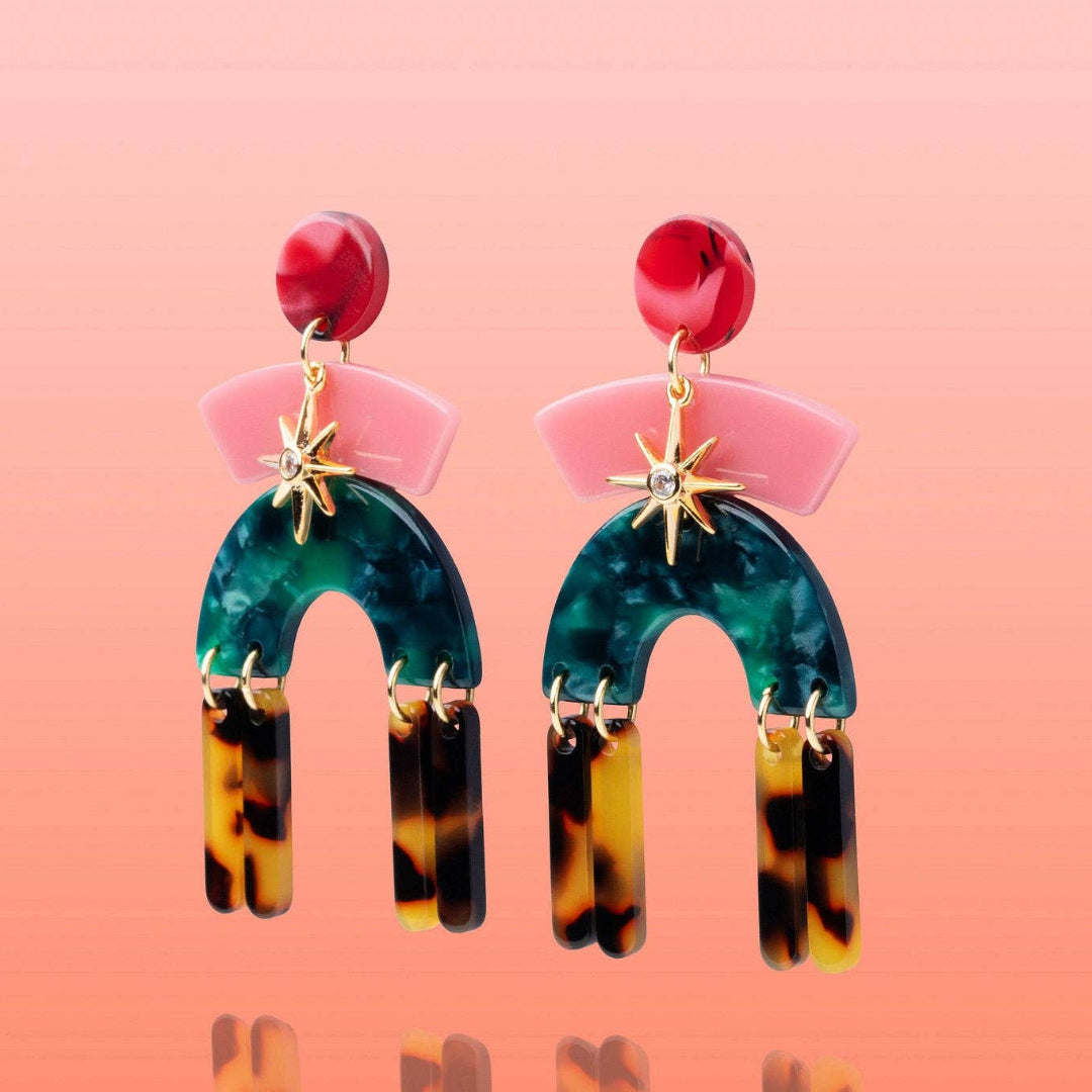 A pair of statement drop earrings with red round studs, pink fan-shaped elements, gold starburst charms, and arch-shaped turquoise and dark green beads hanging from tortoiseshell dangles, set against a peach-pink gradient background.