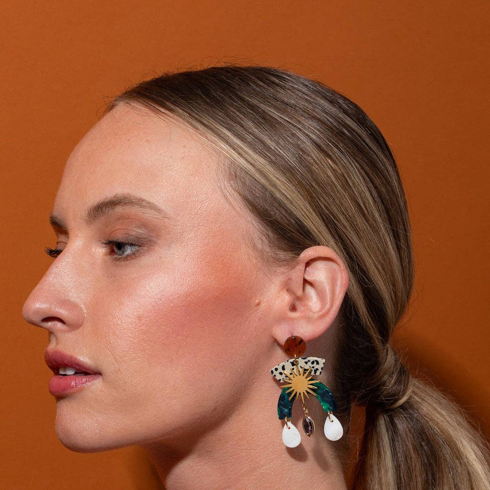 A close-up side profile of a woman with blonde hair in a low ponytail, wearing a colorful statement earring with gold, green, and white details, set against a solid orange background.