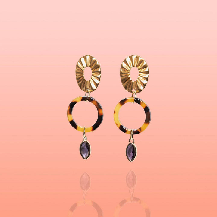 A pair of gold-tone drop earrings featuring a sunburst disc, a tortoiseshell ring, and a dangling marquise-cut purple gemstone, set against a soft pink gradient background.