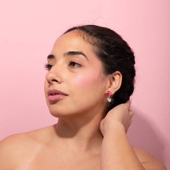 A woman with dark hair pulled back wears a small pink and white flower-shaped earring, posing against a solid pink background.