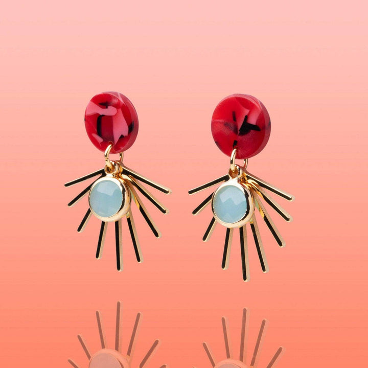 A pair of drop earrings with glossy red circular studs, light blue oval centers, and gold-toned sunburst spikes, shown on a soft peach-pink gradient background.
