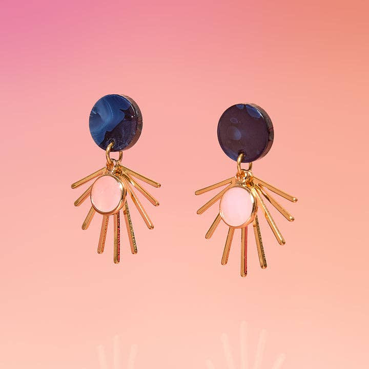 A pair of gold-tone dangle earrings with circular dark blue and pink agate stones, displayed against a soft pink gradient background.