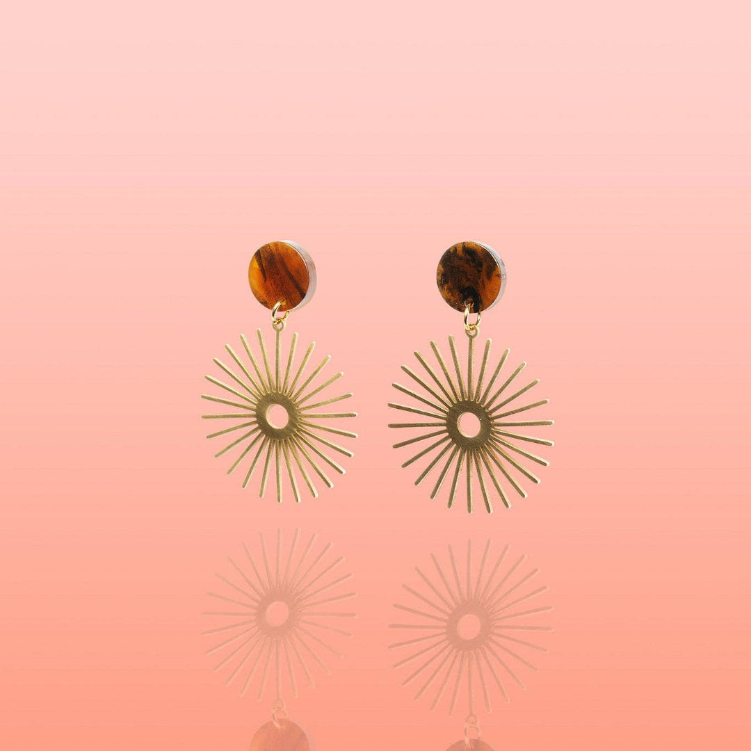 A pair of bohemian-style dangle earrings featuring round brown tortoiseshell studs and gold-toned sunburst disc charms, displayed against a soft pink gradient background with a subtle reflection.