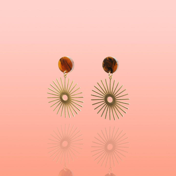 A pair of bohemian-style dangle earrings featuring round brown tortoiseshell studs and gold-toned sunburst disc charms, displayed against a soft pink gradient background with a subtle reflection.