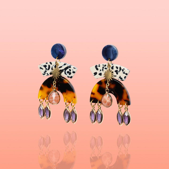 A pair of statement drop earrings on a pink gradient background. Each earring features a dark blue round stud, a white top section with black spots, a gold sunburst charm, a tortoiseshell arch, a pink faceted bead, and three dangling purple teardrop crystals.