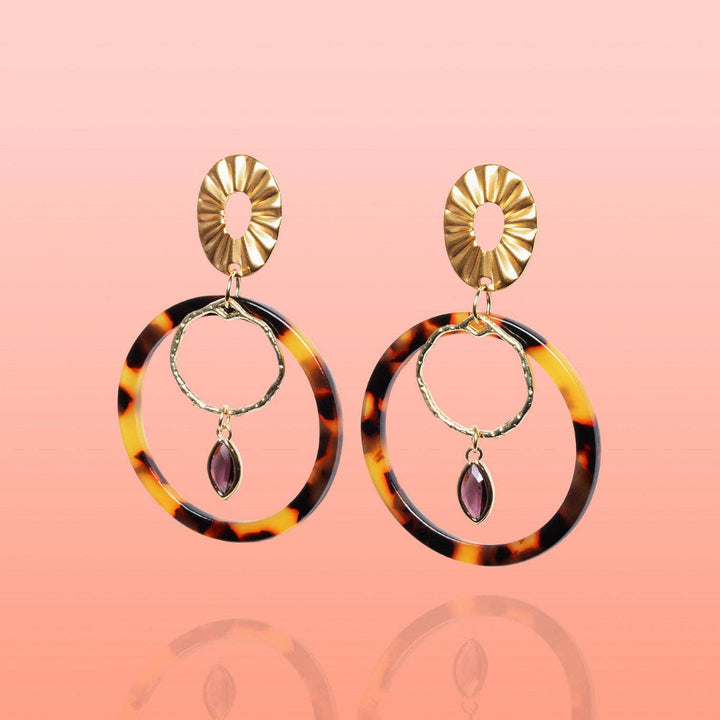 A pair of gold-tone drop earrings featuring a sunburst oval top, a large tortoiseshell circular hoop, a smaller gold ring, and a dangling teardrop-shaped purple gemstone, set against a soft pink gradient background.
