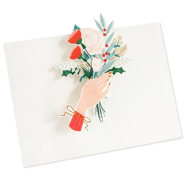 A square white card featuring a layered 3D illustration of a hand holding a bouquet of red strawberries, white roses, green holly, and eucalyptus leaves, set against a clean white background.