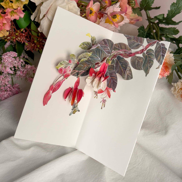 Floral card with red flowers and green leaves on a white surface with flowers in the background