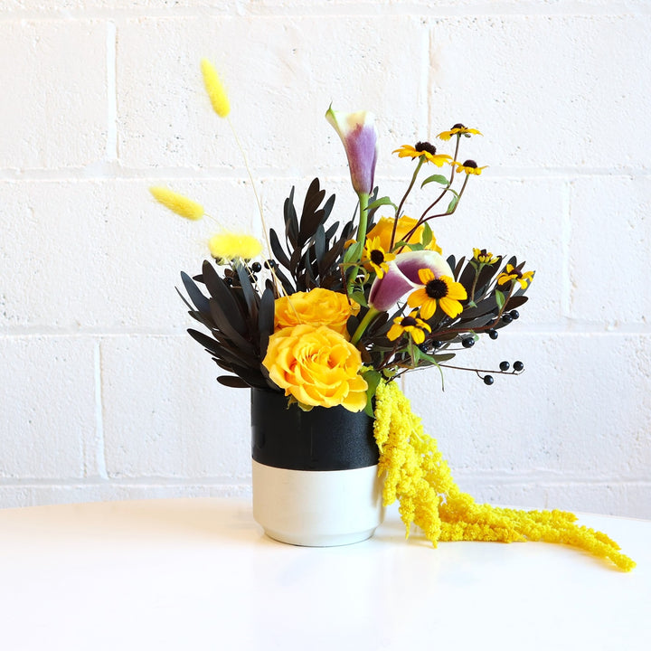 Vibrant | A yellow and black arrangement with a purple accent. Some florals shown are yellow roses, yellow ameranthus, and purple calla lillies. Made in a vase that is black on top and white on the bottom. Photo taken against a white background.