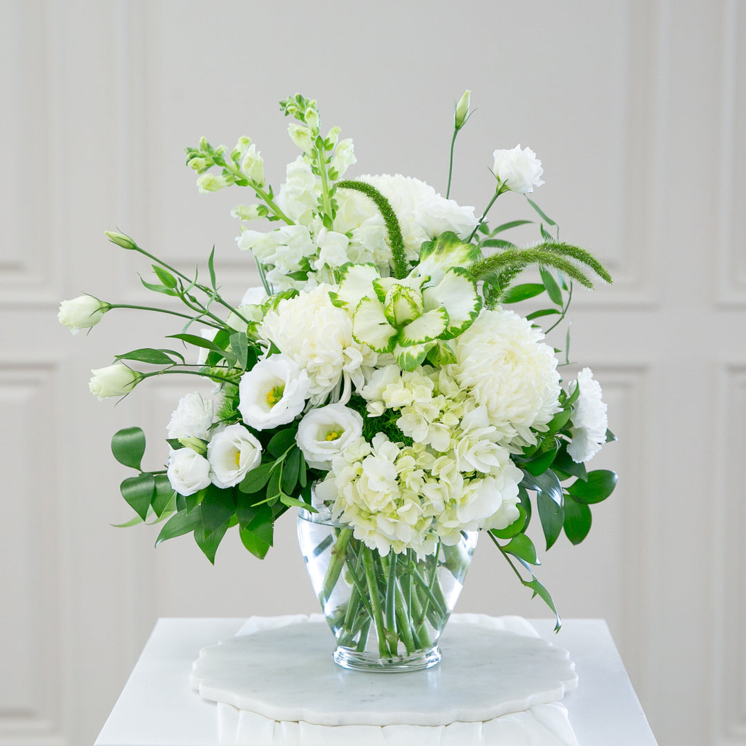 Bouquet of white flowers in a clear vase on a white surface with a light gray wall background