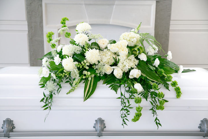 White flowers and greenery on a white casket in front of a gray wall.