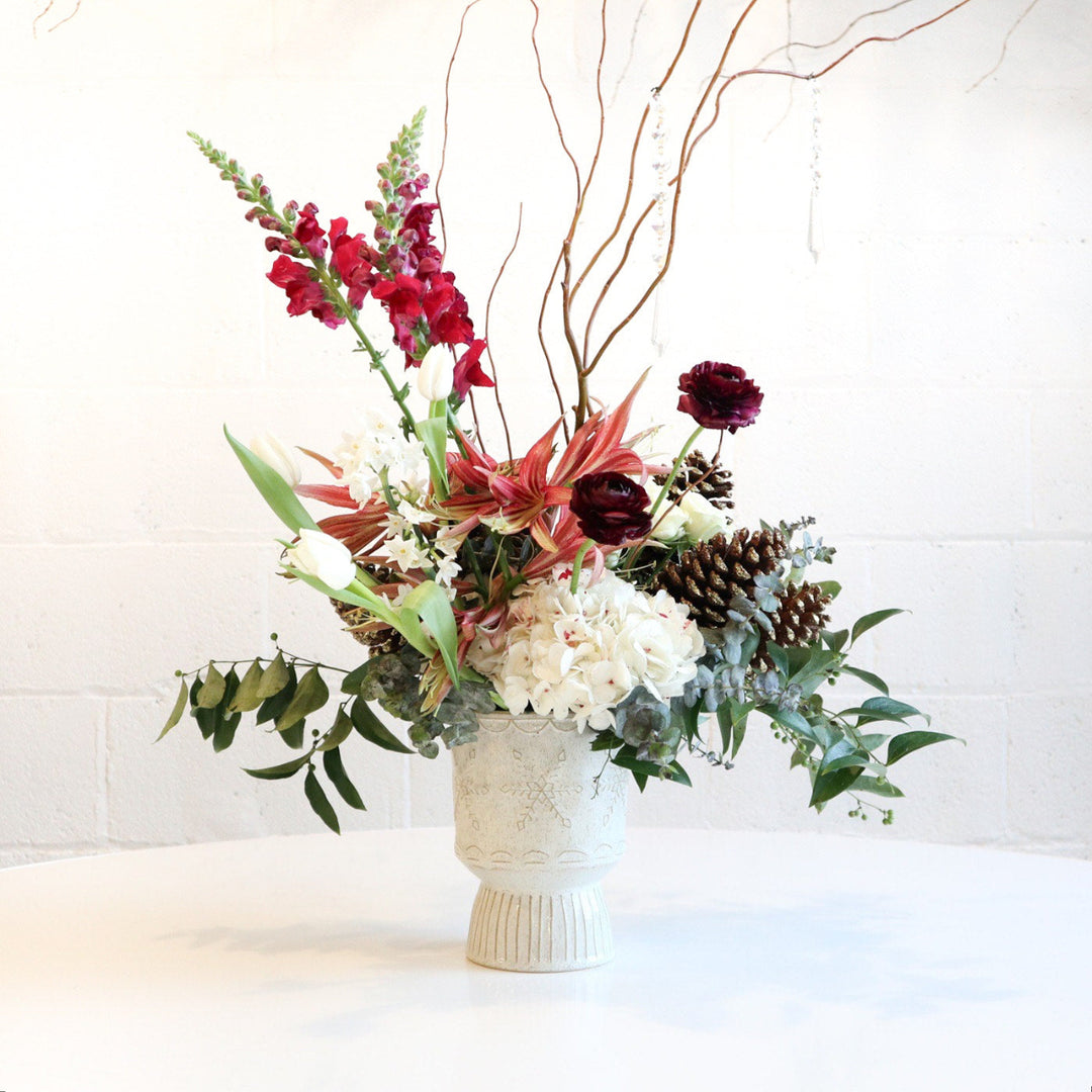 A winter floral centerpiece with a variety of flowers and greenery in a white vase, decorated with pine cones and branches to represent a snow-kissed landscape.