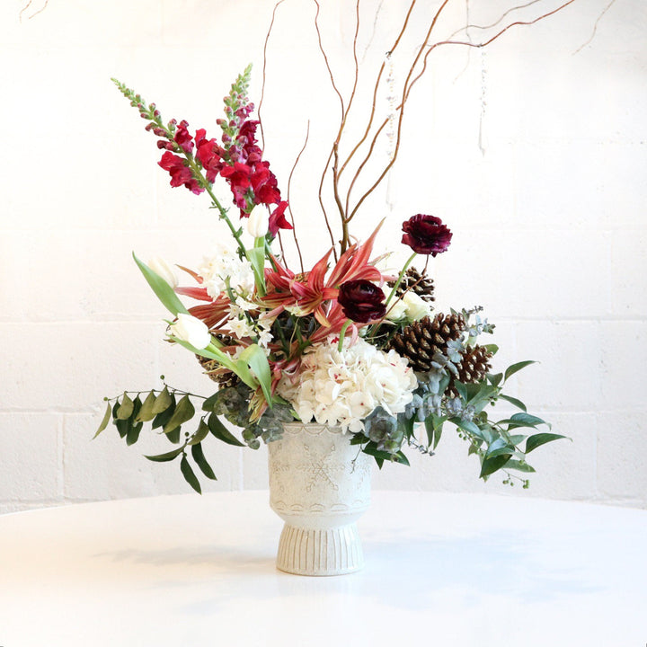 A winter floral centerpiece with a variety of flowers and greenery in a white vase, decorated with pine cones and branches to represent a snow-kissed landscape.
