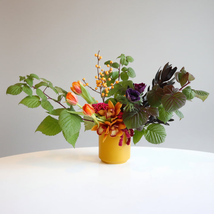 Wonderous | A bright arrangement with orange tulips, orchids, raspberry branches, and other florals in a yellow vase. Photo taken against a white background.
