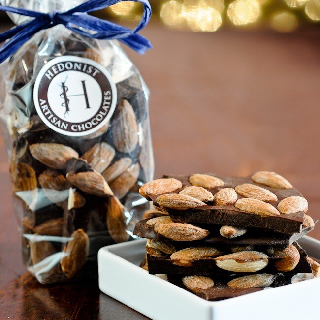 Hedonist Chocolate Bark | Almond Bark, displayed in a white dish with a bag of bark in the background, tied with a blue bow.