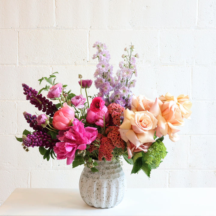 Magnificent | A bright arrangement with the colors purple, pink, peach, and green going from left to right. In a white speckled vase. Photo taken against a white background.