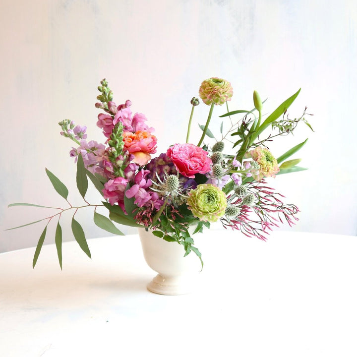 Enchanted | A bright garden arrangement with pinks, purples, and greens in a white vase. Some flowers shown are snapdragons, hydrangea, thistle, and more.
