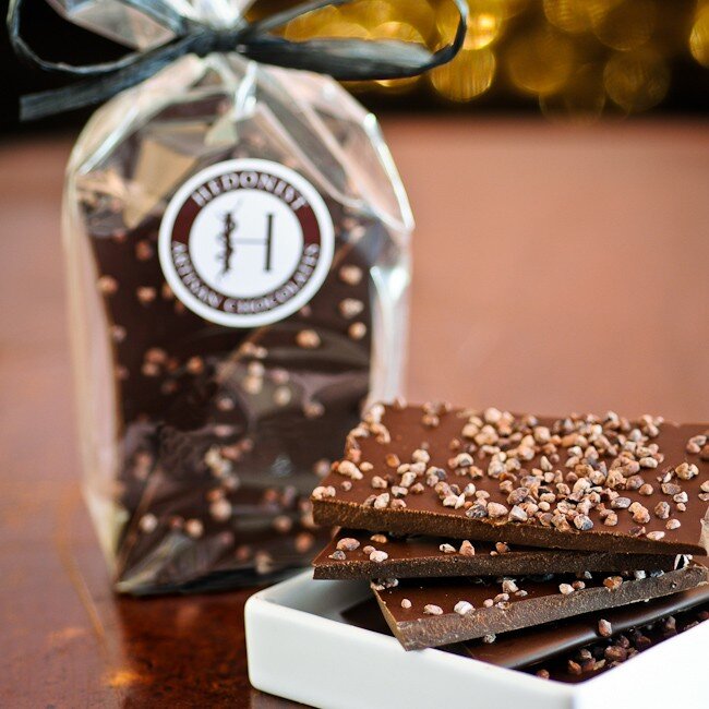 Hedonist Chocolate Bark | Extra Bittersweet Bark, bark displayed in a white dish with more bark on a clear bag in the background tied with a black bow.