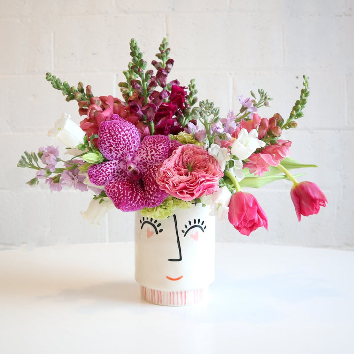 Face vase, with heart eyes a nose and a smile. Filled in the vase is burgundy snapdragons, pink tulips, light purple stock, white campanula and fushia orchid on a white background. 